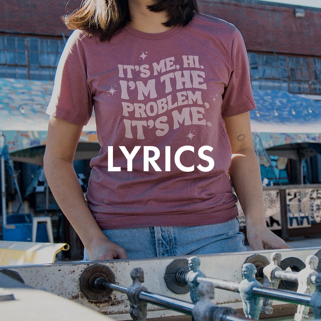 Person wearing a shirt with the text "It's me, hi. I'm the problem, it's me" while standing near a foosball table.