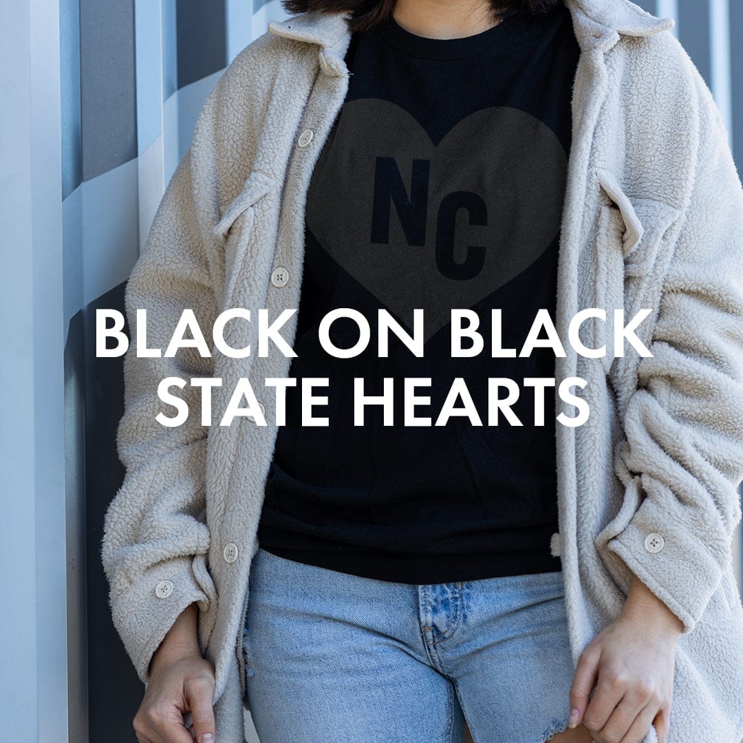 Person wearing The Home T black on black state pride shirt with NC heart design, showcasing state love and soft apparel.